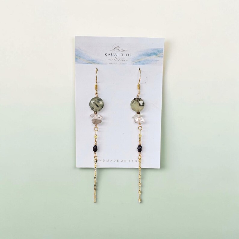 May include: A pair of gold-tone dangle earrings on a white display card labeled Kauai Tide Atelier. Each earring features a faceted sage green bead, a clear crystal chip, a small dark bead, and a delicate gold-tone chain hanging from a hook.