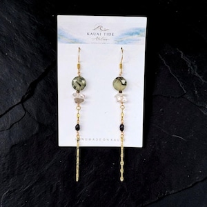 May include: Dangle earrings on a white Kauai Tide Atelier display card featuring faceted sage green round beads, clear crystal chips, small black beads, and long gold-tone textured metal chains hanging from gold-tone ear wires.