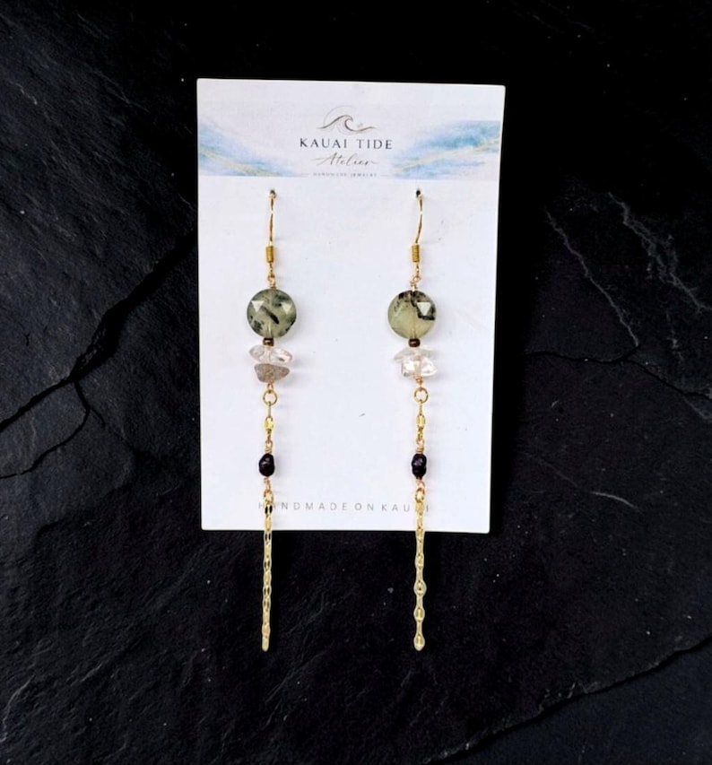 May include: Dangle earrings on a white Kauai Tide Atelier display card featuring faceted sage green round beads, clear crystal chips, small black beads, and long gold-tone textured metal chains hanging from gold-tone ear wires.