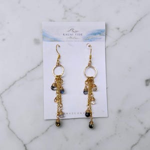 May include: Gold-toned dangle earrings with a circular top and cascading chains. Each chain features a mix of clear, blue, and black teardrop-shaped beads. The earrings hang from a white card with the text "KAUAI TIDE Atelier".