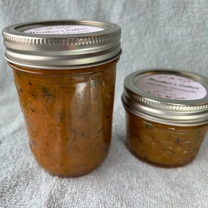 May include: Two glass jars filled with a light orange colored jam. The jars have silver lids and labels that say "Heather's Cellar" with different flavors. The smaller jar says "Mango Pineapple" and the larger jar says "Strawberry Pineapple".