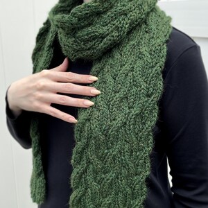 May include: A forest green knitted scarf featuring a textured cable knit pattern. The scarf is draped around the neck and hangs down the front, showcasing the thick yarn construction and the intricate, repeating vertical braided design elements.