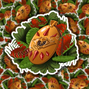 Puede incluir: Un colgante de color dorado con gemas rojas y un diseño detallado, rodeado de follaje verde y rojo. El colgante es una pegatina con un borde blanco.