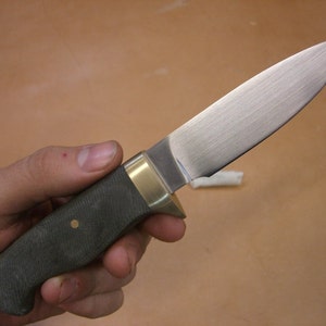 May include: A hunting knife with a dark green handle and a brass bolster. The blade is made of stainless steel and has a satin finish.