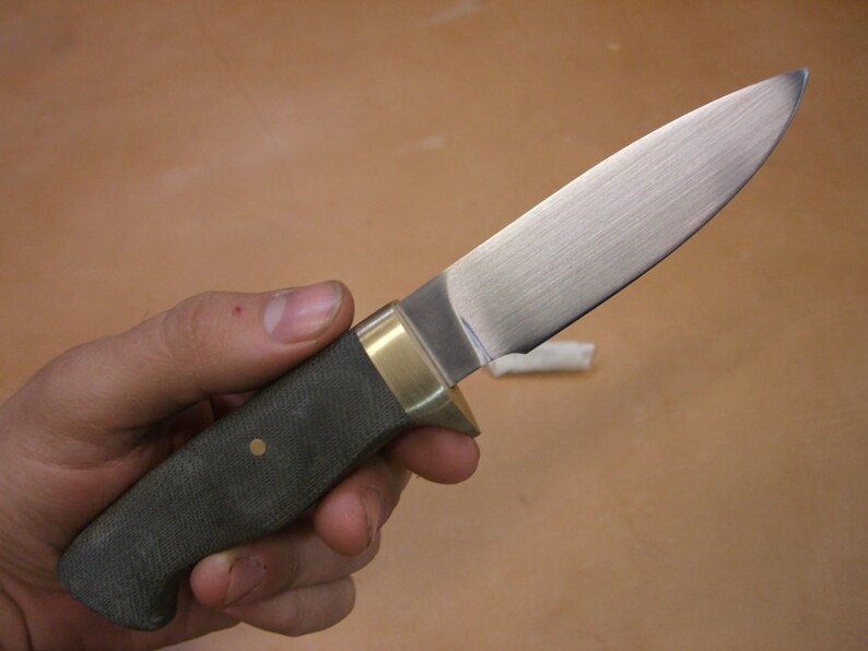 May include: A hunting knife with a dark green handle and a brass bolster. The blade is made of stainless steel and has a satin finish.