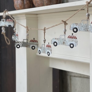 May include: A rustic garland featuring metal trucks carrying pumpkins. The trucks are strung on twine with wooden beads. Two decorative pumpkins sit on a shelf above the garland. The garland is a fall-themed decoration.