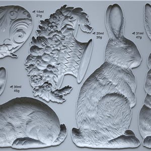 May include: A gray silicone mold featuring various rabbit designs and decorative floral elements. The mold includes rabbit profiles, a seated rabbit, and ornamental pieces. Measurements are indicated in milliliters and grams.