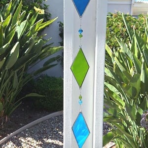 May include: A decorative hanging stained glass suncatcher with three diamond-shaped panels in blue and green. The panels are connected by a silver chain with blue and green beads, and a crystal at the bottom. The suncatcher is hanging outside.