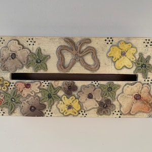 May include: Rectangular wooden box with a cream-colored finish, adorned with embossed floral designs in various colors. A bow-shaped embellishment sits above a slot. The edges are dotted with small black circles. A decorative storage item.
