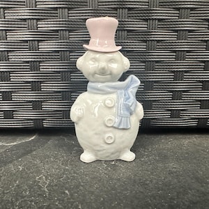 May include: A ceramic snowman figure wearing a light pink top hat and a blue scarf. The snowman has three buttons and a smiling face. The figurine is white and stands on a dark surface.
