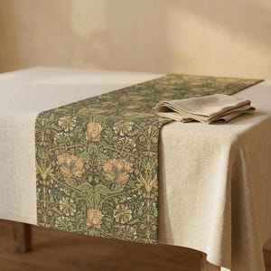 May include: A floral patterned table runner with a matching folded napkin. The runner features a dark green background with a repeating pattern of flowers in shades of tan and green. The napkin is a solid tan color.