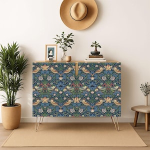 May include: A rectangular wooden cabinet featuring a dark blue floral and bird pattern on its two front doors, supported by four thin gold-toned metal hairpin legs. The cabinet surface holds a small framed picture, a green plant, and books.
