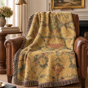 May include: A woven throw blanket featuring a repeating botanical pattern in shades of mustard yellow, sage green, and muted terracotta. The textile has a textured surface and is finished with decorative fringe along the edges, draped over a chair.