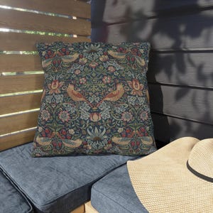 Puede incluir: Cojín decorativo cuadrado con un fondo azul oscuro y un estampado floral y de pájaros en tonos rojos, verdes y beige. El cojín está colocado sobre un cojín azul, con un sombrero de paja en primer plano.