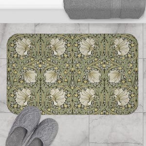 Op de afbeelding: Een rechthoekige badmat met een herhalend bloemmotief in crème, groen en geel. Het ontwerp toont gestileerde bloemen en bladeren op een donkere achtergrond. Grijze pantoffels zijn zichtbaar in de linker benedenhoek.