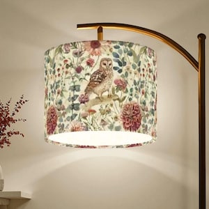 May include: A floor lamp with a curved bronze-toned metal stand and a cylindrical fabric lampshade. The shade features a detailed botanical print with various flowers, green foliage, and a central brown and white owl perched on a branch.