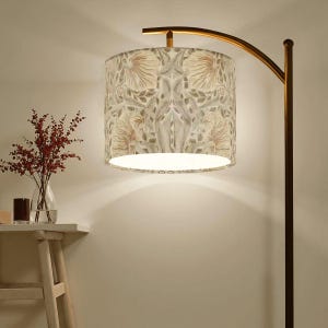 May include: A floor lamp with a curved, gold-toned arm supporting a cylindrical lampshade. The lampshade features a floral pattern in muted tones of beige, pink, and green. A vase of red flowers sits on a small wooden table.
