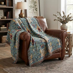 Op de afbeelding: Een geweven plaid met een dicht, ingewikkeld botanisch patroon in tinten blauwgroen, dieprood en gedempt goud. Het textiel heeft zichtbare franjes langs de randen en is gedrapeerd over een bruine leren fauteuil, wat het getextureerde weefsel toont.