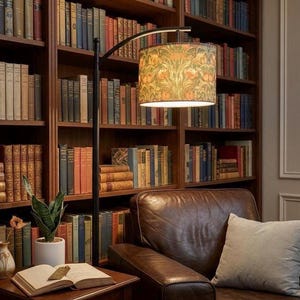 May include: A floor lamp with a thin black metal arched stand and a cylindrical drum lampshade featuring a repeating botanical pattern in shades of green, orange, and yellow, providing warm illumination over a wooden side table and chair.