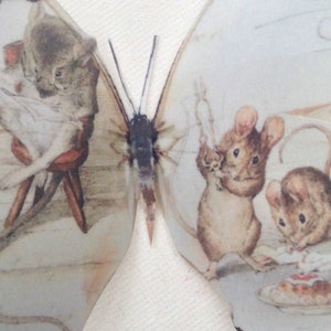 Pode incluir: Um par de asas de borboleta com uma ilustração caprichosa de um rato lendo um livro e dois ratos tomando chá e bolo.
