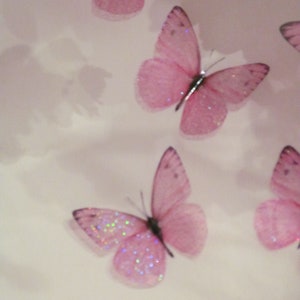 May include: Four pink glitter butterflies with translucent wings are arranged on a white background. The butterflies are different sizes and are positioned in a staggered pattern.