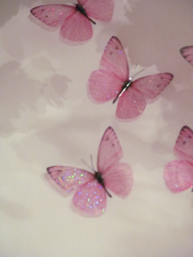 May include: Four pink glitter butterflies with translucent wings are arranged on a white background. The butterflies are different sizes and are positioned in a staggered pattern.