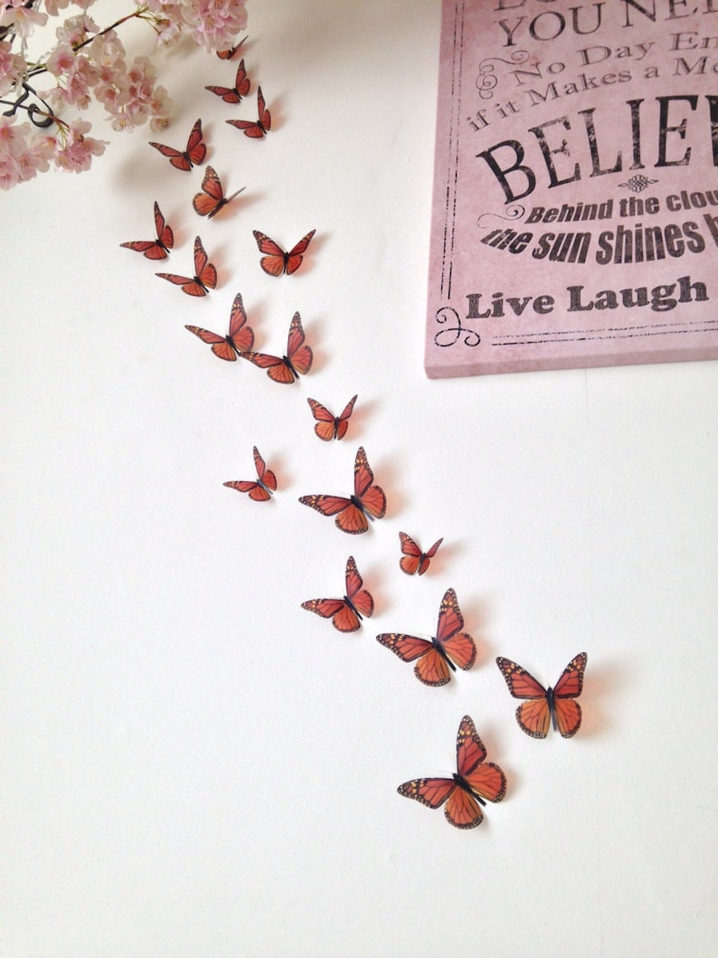 May include: A white wall with a pink sign that reads "You Never Know What a Day Can Bring If It Makes a Moment Believe Behind the Clouds The Sun Shines Live Laugh". A line of orange butterflies are flying up the wall.