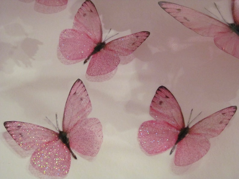 May include: Five pink glitter butterflies with translucent wings. The butterflies are arranged in a scattered pattern on a white background.