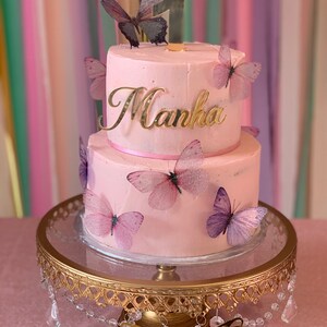 May include: A two-tiered pink cake decorated with purple and pink butterflies. The cake is topped with a gold number one and the name "Manha" in gold lettering.