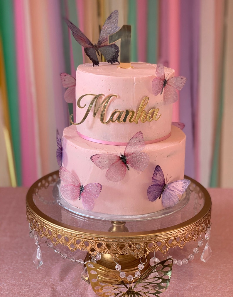 May include: A two-tiered pink cake decorated with purple and pink butterflies. The cake is topped with a gold number one and the name "Manha" in gold lettering.
