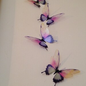 May include: Four iridescent butterflies with purple and pink wings are arranged vertically on a white wall. The butterflies are made of a translucent material and have a delicate, ethereal appearance.