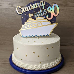 May include: A white frosted cake with gold star sprinkles and a blue base. A cake topper reads "Cruising into 30" with a cruise ship design. The cake is a celebratory dessert.