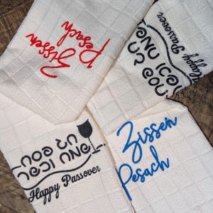 May include: Four white kitchen towels with a textured diamond pattern. Each towel has embroidered text in red, blue, and black, including "Zissen Pesach" and "Happy Passover". The towels are arranged on a wooden surface.