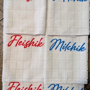 Peut inclure: Six serviettes de cuisine blanches à motifs carrés. Trois serviettes portent le mot "Fleishik" brodé en script rouge, et les trois autres "Milchik" en script bleu. Les serviettes sont pliées et empilées.