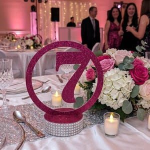 May include: A circular table number sign featuring the number seven in a dark pink glitter finish, mounted on a cylindrical base decorated with rows of silver rhinestones, positioned on a white tablecloth next to a floral centerpiece.