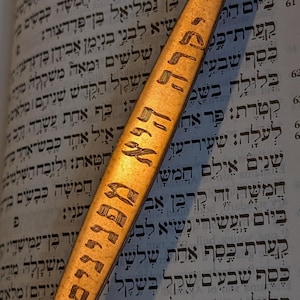 May include: A wooden bookmark with Hebrew text, illuminated against a book's pages. The bookmark is long and slender, with the text etched into its surface. The background is a page from a book with Hebrew script.