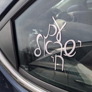 May include: A white vinyl decal applied to a car window featuring stylized Hebrew calligraphy. The design consists of three stacked lines of characters in a flowing, modern script, creating a vertical arrangement on the glass surface.