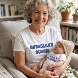 May include: A white t-shirt with the blue text BUBBELEH'S BUBBIE and a white baby bodysuit with the blue text BUBBIE'S BUBBELEH. The baby wears a tan knit beanie. The clothing items are displayed together, featuring matching graphic text designs.