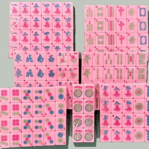 Op de afbeelding: Roze Mahjong-tegelsets, elk verpakt in doorzichtig plastic. De tegels tonen verschillende symbolen, cijfers en Chinese karakters in blauw, groen en rood. Een klassiek spel.