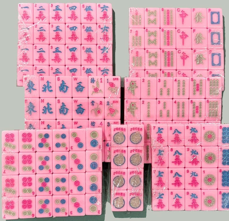 Op de afbeelding: Roze Mahjong-tegelsets, elk verpakt in doorzichtig plastic. De tegels tonen verschillende symbolen, cijfers en Chinese karakters in blauw, groen en rood. Een klassiek spel.