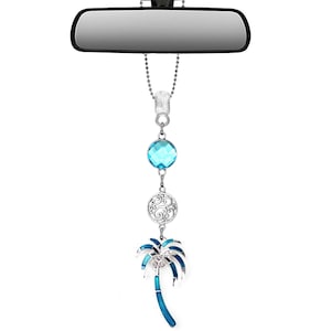 May include: A car charm hanging from a rearview mirror. The charm features a faceted blue crystal, a silver filigree disc, and a blue and silver palm tree design. The charm is suspended by a silver chain.