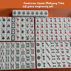 Op de afbeelding: Een complete set American Home Mahjong-tegels. De 152-delige graveerset bevat witte tegels met zwarte, rode en groene symbolen en karakters. De set bevat Jokers, winden, draken en suit-tegels. De tegels zijn gerangschikt op een oranje oppervlak.