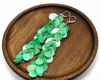 May include: A pair of long dangle earrings featuring multiple layers of circular, light green, iridescent sequins attached to silver-toned metal lever-back closures. The sequins have a smooth, reflective finish that catches the light.