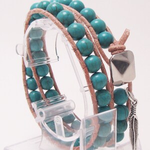May include: A turquoise and brown leather wrap bracelet with a feather charm. The bracelet is made with turquoise beads and brown leather cord. The feather charm is silver and has a turquoise bead at the top.