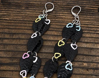 May include: A pair of dangle earrings featuring silver-toned leverback hooks, black leaf-shaped charms, and small pastel-colored heart-shaped links in yellow, pink, and light blue, arranged in a cascading vertical design with metal jump rings.