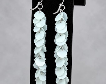 May include: A pair of long dangle earrings featuring multiple pale mint green circular discs attached to silver-toned metal hooks. The discs are layered in a cascading vertical arrangement, creating a textured, fringe-like appearance that hangs down from the top ear wire.