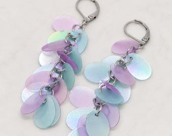 May include: A pair of dangle earrings featuring multiple iridescent, oval-shaped sequins in shades of pastel purple and light blue. The sequins are attached to silver-toned metal jump rings and suspended from silver-toned lever-back earring hooks.
