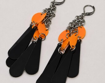 May include: A pair of dangle earrings featuring dark metal lever-back closures, silver-toned chain links, and a cluster of dangling elements. The design includes several bright orange circular discs and elongated black teardrop-shaped pendants.