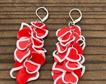 May include: A pair of dangle earrings featuring a cluster of layered heart-shaped charms in vibrant red and white. Each earring is suspended from a silver-toned metal leverback clasp, creating a cascading effect with multiple overlapping heart cutouts.