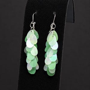 May include: A pair of dangle earrings featuring multiple overlapping, teardrop-shaped sequins in shades of light green and iridescent white. The sequins are attached to silver-toned metal hooks, creating a layered, cascading effect that hangs vertically.
